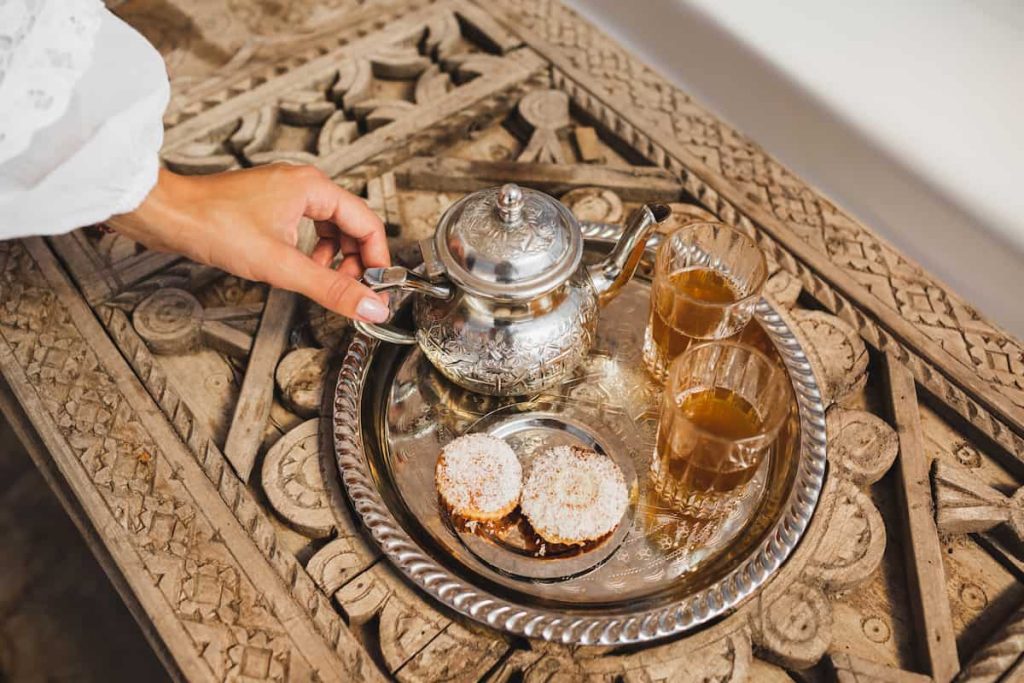 Woman hands serving traditional moroccan mint tea ceremony with cookies and vintage silver teapot. 
