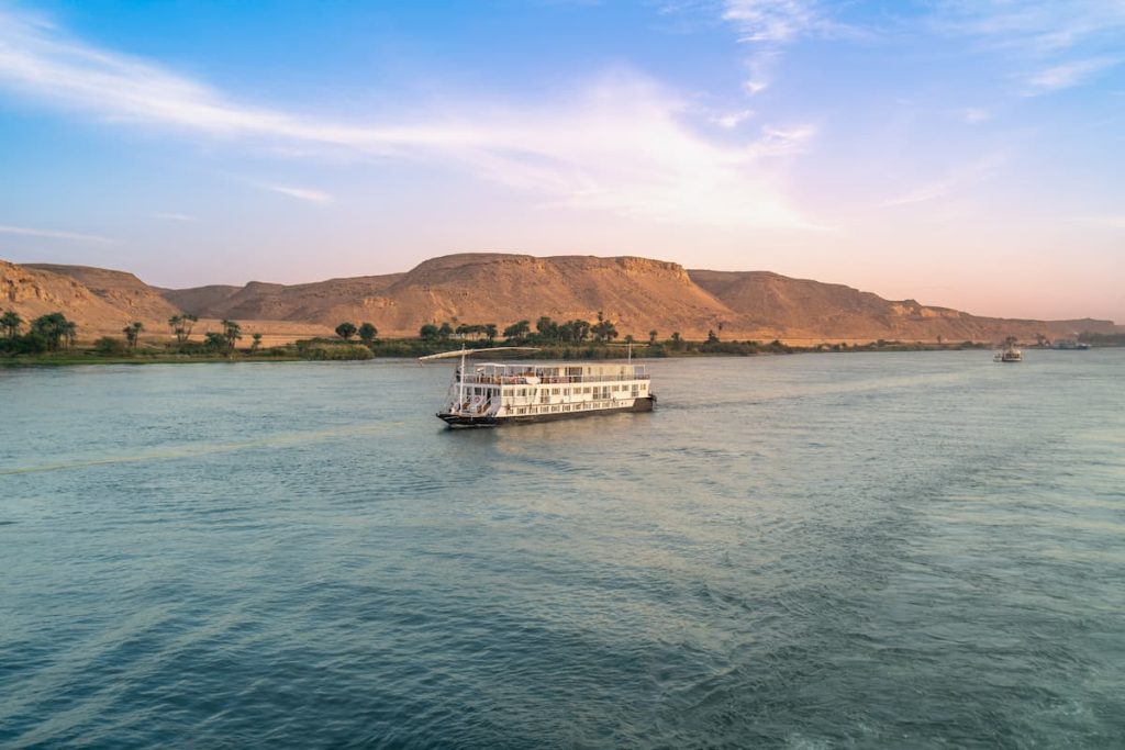 A tranquil scene on the Nile River near Esna Lock in Aswan, Egypt, as two cruise boats navigate the waters during a beautiful dusk with soft, colorful skies and distant desert banks.