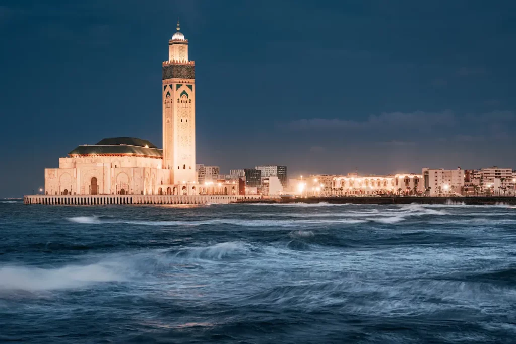 Hassan II Mosque in Casablanca, Morocco, radiating with a warm glow at dusk, while gentle waves of the Atlantic Ocean lap at the shore