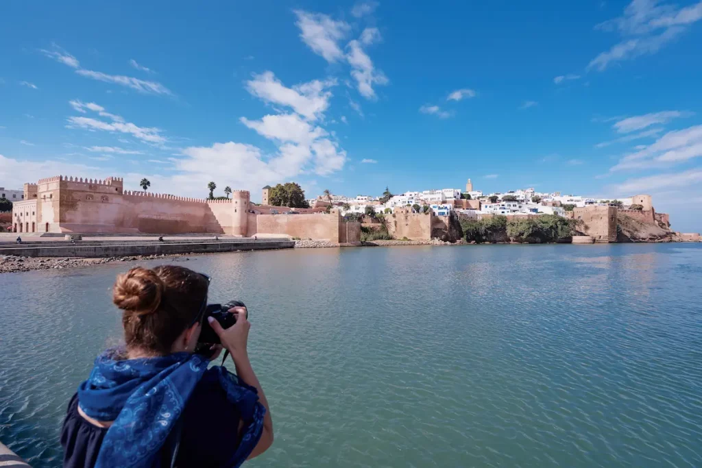 A woman photographs the historic Kasbah of the Udayas in Rabat, Morocco, with a clear blue sky and calm river in the foreground.