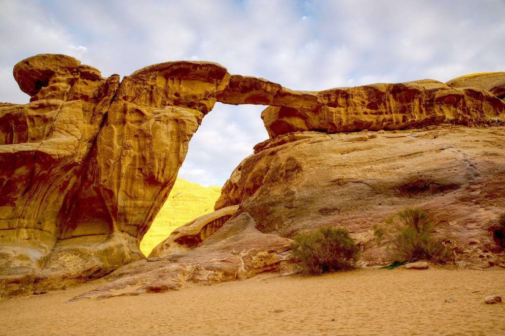 Wadi Rum Burdah Rock Bridge in Jordan with cloudy sky and golden-colored mountain in background Amazing Stone Rock Arch in Jordan, Wadi Rum desert